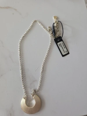 Silverfärgat halsband med rund hängare - Snyggt halsband i silverfärg med en rund, halvmåneformad hängare. Kedjan är tunn och elegant, perfekt för att lyfta vilken outfit som helst. Hängaren har en slät, blank yta och halsbandet har en enkel men modern design.