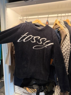 Mörkblå fluffig stickad tröja - DEN ÄR LITE OVERSIZE. PASSAR M BÄST! Mysig mörkblå stickad tröja med fluffig textur och vit texten 'tossy' i stor skrivstil över bröstet. Tröjan har rund halsringning och långa ärmar, perfekt för kyligare dagar och en avslappnad stil.
