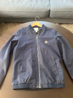 Marinblå bomberjacka från Moncler - Snygg marinblå bomberjacka från Moncler med klassisk zip framtill och ribbade muddar. Jackan har två sidofickor och Moncler-logga på bröstet. Perfekt för dig som vill ha en stilren och trendig look.