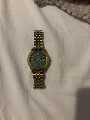 Rolle - Snygg klocka med länkarmband i silver och guldton, samt grön urtavla med tydliga markeringar och datumvisning. Boetten är rund och har en guldfärgad ring runt urtavlan. Perfekt accessoar för dig som gillar stilrena detaljer.