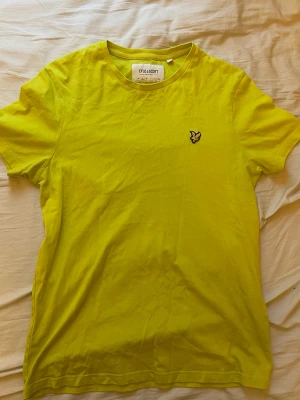 Gul t-shirt från Lyle & Scott - Fräsch gul t-shirt från Lyle & Scott med klassisk rund hals och broderad logga på bröstet. Tillverkad i mjuk bomull och har en enkel, clean design som passar perfekt till jeans eller shorts.