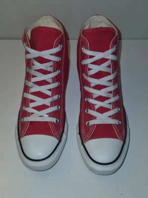 Röda Converse Chuck Taylor sneakers - Klassiska röda Converse Chuck Taylor All Star high-top sneakers med vit gummisula och tå, vita skosnören och metallöljetter. Skorna är i canvasmaterial och har den ikoniwska Converse-loggan på sidan. Perfekta för en cool och avslappnad stil.