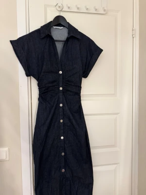 Mörkblå jeansklänning med knappar från Zara  - Snygg mörkblå jeansklänning med krage och korta ärmar. Klänningen har knappar längs hela framsidan och en markerad midja med rynkdetaljer. Perfekt för dig som gillar en avslappnad men ändå stilren look. Bara använt 1 gång. 