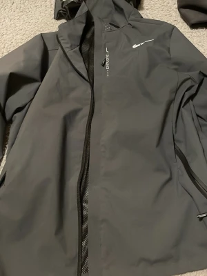 Svart regnjacka från Nike med huva - Snygg svart regnjacka från Nike med huva och dragkedja. Jackan har meshfoder på insidan och är vatten- och vindtät. Diskret Nike-logga på bröstet och praktiska detaljer för extra skydd mot väder. Perfekt för regniga dagar.