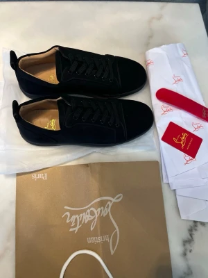 Svarta Christian Louboutins - Stilrena svarta sneakers i mocka från Christian Louboutin med klassisk röd sula och svarta skosnören. Låg modell med diskret logga på innersulan och lyxiga detaljer. Perfekta för dig som vill ha en exklusiv touch på din outfit.