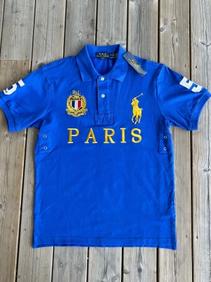 Chief Keef ”Paris” pikétröja från Polo Ralph Lauren. - Snygg blå pikéskjorta från Polo Ralph Lauren med gul broderad logga och texten PARIS framtill. Skjortan har klassisk krage, korta ärmar och vita knappar. Detaljer som broderat märke och siffror på ärmarna ger en sportig look. Perfekt för dig som gillar streetwear. Aldrig använd, inga defekter och har fortfarande prislapp.