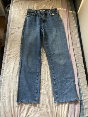 Blå raka jeans med rå kant - Säljer ett par klassiska blå jeans med rak passform och rå, fransig kant längst ner på benen. Jeansen har fem fickor och hög midja. Perfekta för en avslappnad och trendig look. Materialet är kraftig denim i 100% bomull.
