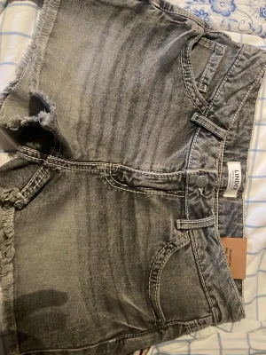 Grå broderade jeansshorts från Lindex - Snygga grå jeansshorts från Lindex med slitna kanter och broderade blommor i blått, gult och vitt på bakfickorna. Klassisk femficksmodell med knapp och dragkedja framtill. Perfekta för en avslappnad sommarstil.