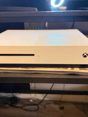 Vit Xbox One S spelkonsol - Säljer en stilren vit Xbox One S med klassisk rektangulär form och ventilerad ovansida. Konsolen har en diskret design med svarta detaljer och är tillverkad i plast. Perfekt för gaming och streaming!