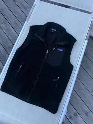 Pantagonia väst - Svart fleeceväst från Patagonia med dragkedja framtill, två fickor med dragkedja och en bröstficka med logga. Västen har hög krage och är gjord i mjukt, teddy-liknande material. Perfekt för lager-på-lager och outdoor-stil.