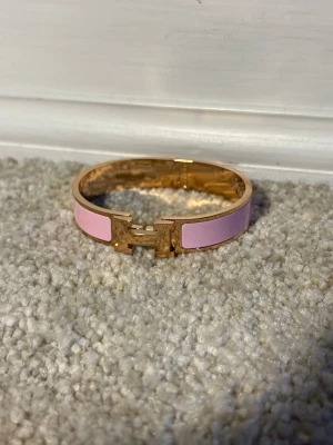 Rosa och guldigt armband med H-detalj - Snyggt armband i guldton med ljusrosa emaljpaneler och en ikonisk H-formad detalj framtill. Armbandet har en stilren och modern design som verkligen sticker ut. Perfekt accessoar för att lyfta din outfit med en touch av färg och elegans.