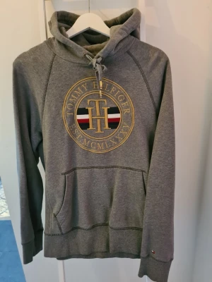 Grå hoodie från Tommy Hilfiger - Snygg grå hoodie med huva och dragsko från Tommy Hilfiger. Stor broderad logga i guld, rött, vitt och blått framtill. Klassisk känguruficka och ribbade muddar. Perfekt för en avslappnad och trendig stil.