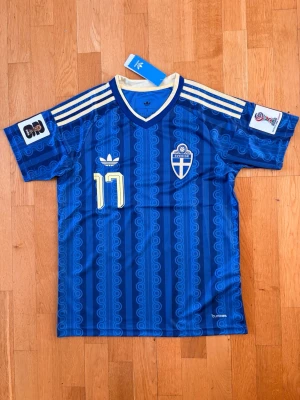 Blå Sverige fotbollströja nr 17 - Säljer en blå fotbollströja från Adidas med Sveriges landslagsemblem, nummer 17 och namnet Gyökeres på ryggen. Tröjan har vita och gula detaljer, klassiska Adidas-ränder på axlarna och korta ärmar. Perfekt för match eller träning!