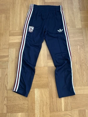 Adidas Arsenal byxor marinblå - Marinblå  sköna byxor från Adidas x Arsenal med vita och vinröda ränder längs sidorna. Byxorna har Arsenal-logga och Adidas Trefoil på framsidan samt elastisk midja. Perfekta för sport eller chill. 