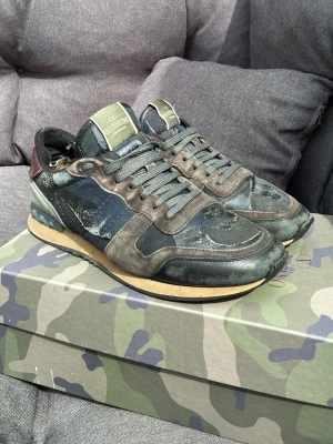 Valentino camo sneakers i grönt - Snygga sneakers från Valentino med camomönster i olika nyanser av grönt, svart och beige. Skorna har en klassisk låg siluett, gröna skosnören och detaljer i mocka och läder. Sulan är beige med svarta och blå inslag baktill. Perfekta för dig som gillar streetwear och exklusiv stil. Dom är väl använda därav är priset lite lägre skriv om ni har några frågor