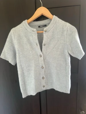 Ljusgrå kortärmad kofta Gina Tricot - Kortärmad kofta från Gina Tricot i en ljusgrå nyans och använd en gång. Koftan har rund halsringning, ribbade kanter och knappar framtill. Perfekt att styla över ett linne eller t-shirt i sommar 🩵