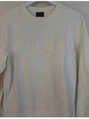 Vit sweatshirt från Oscar Jacobson - Klassisk vit sweatshirt från Oscar Jacobson med diskret broderad logga på bröstet. Tröjan har rund halsringning och ribbade muddar vid ärmslut och nederkant. Perfekt för en clean och stilren look.