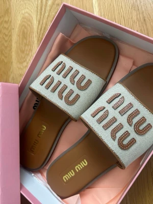 Beige sandaler från Miu Miu - Snygga beige sandaler från Miu Miu med bred rem i canvas och logotyp i brunt broderi. Innersulan är i brunt läder och yttersulan är slät. Kommer i original ask och med dustbag. Storlek 41, men passar mig som har 40