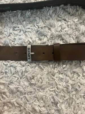 Brunt skärp från Levi's i skinn - Snyggt brunt skärp från Levi's i äkta skinn med klassiskt silverfärgat spänne där Levi's-loggan syns tydligt. Perfekt till jeans eller chinos för en clean och stilren look.