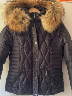 Svart dunjacka med stor pälshuvan som ny - Snygg svart dunjacka med quiltat mönster och stor luva kantad med fluffig päls i bruna toner. Jackan har dragkedjor på fickorna och en extra bröstficka med dragkedja. Perfekt för kalla vinterdagar och ger en riktigt trendig look. Bröst 96cm, längd 62cm. Filmar när jag ska packa och skicka.