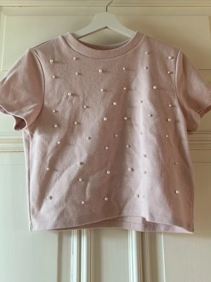 Ljusrosa t-shirt med pärlor H&M Divided - Supersöt ljusrosa t-shirt från H&M Divided, dekorerad med pärlor på framsidan för en extra stylish look. Klassisk rund halsringning och korta ärmar. Perfekt att matcha med jeans eller kjol för en trendig vibe. Materialet är mjukt på utsidan. Använd fåtal gånger