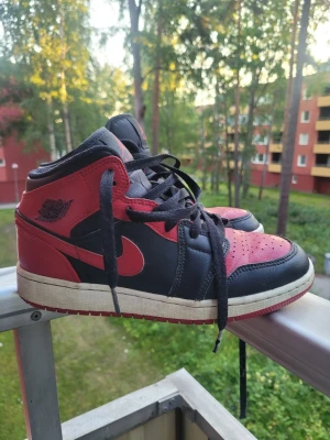 Air Jordan 1 Mid Svart/Röd - Säljer ett par Air Jordan 1 Mid sneakers i svart och rött läder med klassisk röd swoosh och Air Jordan-logga på sidan. Skorna har svart snörning, vit mellansula och röd yttersula. Perfekta för dig som gillar retro och streetwear vibes.
