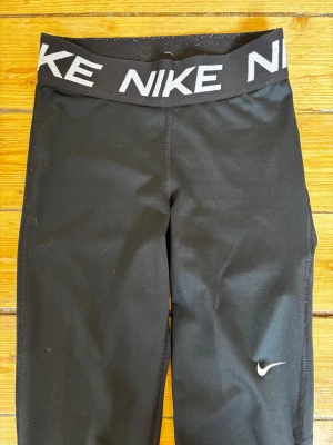 Svarta träningstights från Nike storlek XS - Snygga svarta träningstights från Nike med hög midja och bred resår med stor Nike-logga. Tightsen har en liten vit swoosh på benet och är storlek XS.