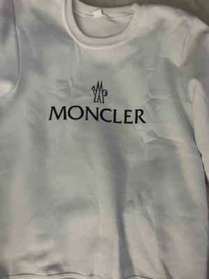 Vit sweatshirt från Moncler - Snygg vit sweatshirt från Moncler med klassisk logga tryckt i svart på bröstet. Tröjan har rund halsringning och ribbade muddar vid ärmslut och nederkant. Perfekt för en clean och stilren look.