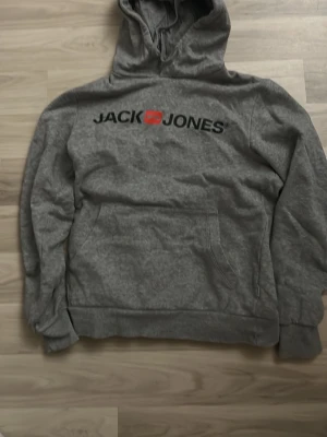 Grå hoodie från Jack & Jones - Säljer en grå hoodie från Jack & Jones med stor logga i svart och rött på bröstet. Hoodien har en klassisk huva med dragsko och en stor magficka. Mjuk insida i fleece och ribbade muddar vid ärmar och nederkant. Perfekt för chill dagar.