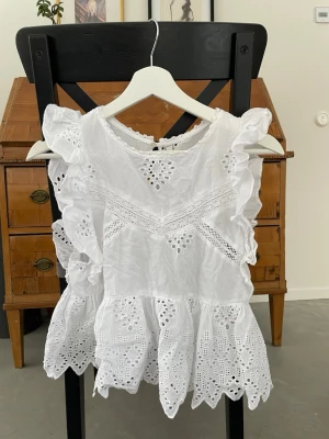 Vit broderad blus från Zara - Superfin vit blus från Zara med broderade detaljer och volanger längs axlarna. Blusen har ärmlös design, spetsmönster och dekorativa hålbroderier. Perfekt för sommarens alla tillfällen och ger en romantisk vibe.