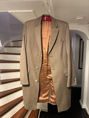 Oscar Jacobson rock - Stilren beige kappa från Oscar Jacobson med tre knappar framtill och slät finish. Fodrad med glansigt tyg i kopparfärg. Klassisk krage, två fickor och dekorativa knappar vid ärmslut. Perfekt för dig som gillar tidlös och elegant stil.