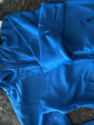 Blå hoodie microfleece  med dragkedja - Snygg blå hoodie från Cross med dragkedja framtill och huva. Tillverkad i ett mjukt och stretchigt material, perfekt för en sportig look. Fickor med dragkedja på sidorna och tumhål vid ärmsluten för extra komfort.