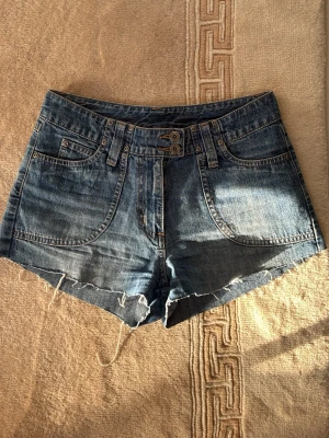 Blå jeansshorts med rå kant - Snygga blå jeansshorts med rå, fransig kant och dubbla knappar i midjan. Perfekta för varma dagar och enkel att matcha med allt.