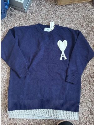 Mörkblå stickad tröja med hjärta - Säljer en mörkblå stickad tröja från AMI Paris med ett vitt hjärta och bokstaven A på bröstet. Tröjan har rund halsringning, långa ärmar och ribbade muddar vid ärmslut och nederkant. Perfekt för dig som gillar stilrena detaljer.