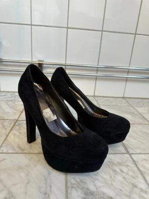 Svarta platåpumps i mocka-look 39 - Säljer ett par klassiska svarta pumps med hög platå och chunky klack. Skorna har rundad tå och är klädda i mockaimitation som ger en lyxig känsla. Perfekta för dig som vill ha extra höjd och stilren look. Lite slitna se sista bild 