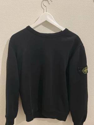 Svart sweatshirt från Stone Island - Säljer en svart sweatshirt från Stone Island med klassisk rund hals och ikonisk patch på vänster ärm. Tröjan har ribbade muddar vid ärmslut och nederkant. Tillverkad i mjuk bomull för skön passform och stilren look. Storlek S. Varken lappen eller kvitto finns kvar. Därav det låga priset. Äkthet kan endast garanteras av mig personligen.