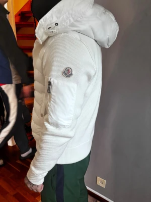 Vit pufferjacka från Moncler - Snygg vit pufferjacka från Moncler med ribbade ärmar och vadderad front. Jackan har en stor huva, ficka med dragkedja på ärmen och klassisk Moncler-logga. Tillverkad i en mix av dun och stickat material för extra värme och stil.