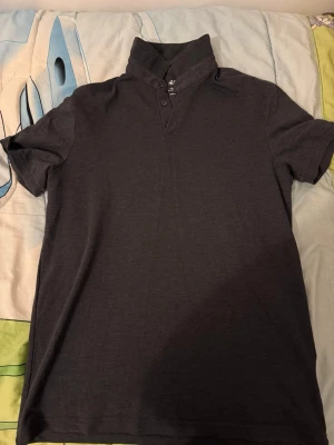 Mörkgrå polotopp från H&M - En stilren mörkgrå polotopp från H&M med korta ärmar och knappar vid halsen. Toppen har en enkel design och är tillverkad i mjukt material som känns skönt mot huden. Perfekt för en avslappnad och trendig look.