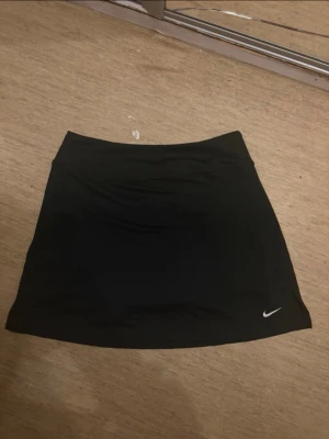 Svart skort från Nike - Snygg svart skort från Nike med klassisk vit logga broderad längst ner. Skorten har en enkel och sportig design med rak passform och elastisk midja. Perfekt för dig som gillar en clean och stilren look. Materialet är mjukt och stretchigt för maximal komfort.