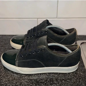 Lanvin svarta sneakers mocka/lack - Snygga svarta sneakers från Lanvin med ovandel i mocka och lackad tå. Skorna har vita, platta sulor och svarta snören. Klassisk och stilren design med diskreta sömmar och metallöljetter. Perfekta för dig som gillar lyxiga och minimalistiska sneakers. 