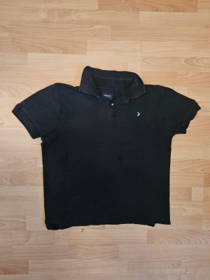 Svart pikétröja med broderad logga - Klassisk svart pikétröja från Boomerang med korta ärmar och krage. Tröjan har en liten broderad logga framtill i grönt och orange. Tillverkad i bomull för en bekväm känsla och stilren look.