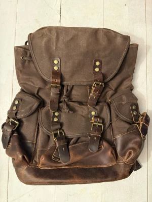 Brun vintage retro ryggsäck i canvas och läder - Snygg brun ryggsäck i kraftig canvas med detaljer i mörkbrunt läder. Flera spännen i metall och läderremmar ger en retro känsla. Ryggsäcken har flera ytterfickor och kontrastsömmar. Perfekt för dig som gillar klassisk och robust stil. Helt ny! Ny på Amazon 3030kr. 