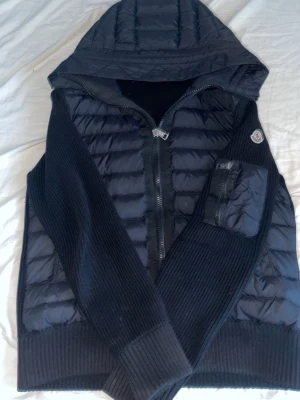 Svart pufferjacka med huva från Moncler - Snygg svart pufferjacka från Moncler med stickade ärmar och huva. Jackan har dragkedja framtill, en ficka på ärmen och klassisk Moncler-logga på vänster ärm. Materialmix av quiltad nylon och ribbstickad ull ger både värme och stil.