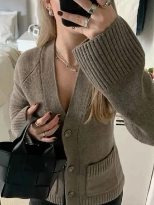 Beige stickad kofta med knappar - Mysig beige stickad kofta med stora knappar framtill och breda ribbade muddar vid ärmsluten. Koftan har en djup V-ringning och två fickor framtill. Perfekt för lager på lager och en avslappnad stil.