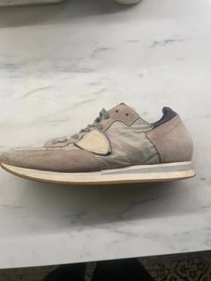 Beige sneakers från Philippe Model - Snygga beige sneakers från Philippe Model med gråa skosnören, mockadetaljer och textilpaneler. Skon har en ikonisk sköldlogga på sidan och vit sula med beige kant. Perfekt för dig som gillar stilrena och klassiska sneakers.