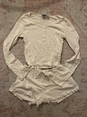 Vit ribbad pyjamas från Gina Tricot - Supersöt vit pyjamas från Gina Tricot i ribbat material. Setet består av en långärmad topp med knappar framtill och volangkant samt matchande shorts med dragsko och volangdetaljer. Perfekt för mysiga hemmakvällar.