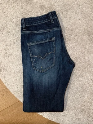 Mörkblå slitna jeans från Guess - Mörkblå jeans från Guess med raka ben, slitningar och distressed detaljer framtill. Klassisk femficksmodell med kontrastsömmar och snygg bakficka med dekorativ söm. Perfekta för en avslappnad och trendig look.