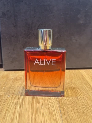Alive parfym med guldlock - Alive parfym från Hugo Boss i en stilren, rektangulär glasflaska med röd-orange doftvätska och ett elegant guldigt lock. Flaskan har enkel och modern design med texten 'ALIVE' på framsidan. Perfekt för dig som gillar exklusiva dofter.