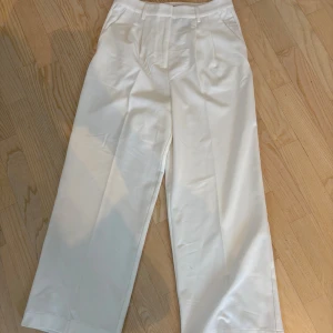 Vida vita kostymbyxor - Säljer ett par vita kostymbyxor med hög midja och vida ben. Byxorna har pressveck framtill, bälteshällor och diskreta fickor. Perfekta för en clean och stilren look.