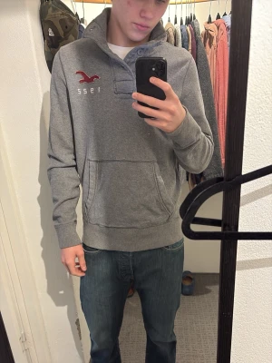 Grå vintage hoodie från Hollister - Snygg grå hoodie från Hollister med broderad röd logga och årtalet 1922 på bröstet. Tröjan har känguruficka framtill, ribbade muddar och knappdetalj vid halsen. Perfekt för en avslappnad och stilren look.storlek M väldigt bra skick. Hör av er vid frågor